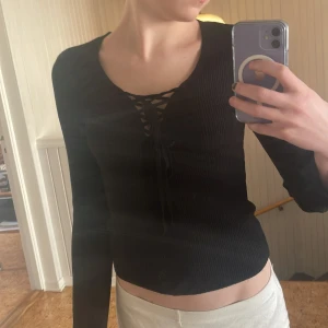 Svart ribbad topp med snörning - Snygg svart ribbad topp med lång ärm och snörning framtill. Perfekt för en stilren look. Passar bra till både jeans och kjol.