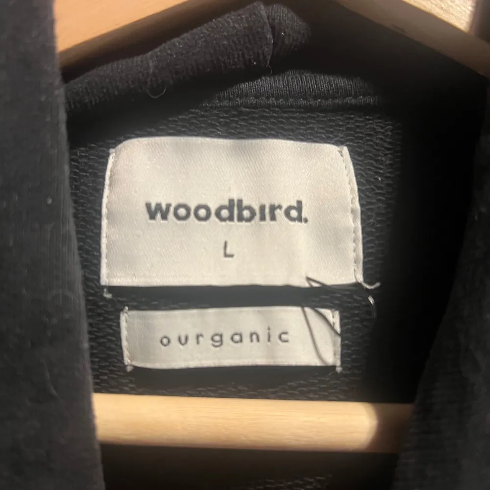 Säljer en svart hoodie från Woodbird med en liten logga på bröstet. Den har en klassisk design med dragsko i huvan och en stor magficka. Perfekt för en avslappnad stil.. Neuletakit & Villapaidat.