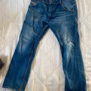 Blå jeans från Diesel - Säljer ett par klassiska blå jeans från Diesel med en cool slitning på ena benet. De har en straight passform och är tillverkade i slitstarkt denim. Perfekta för en avslappnad stil.