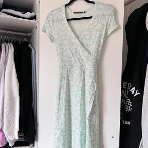 Mintgrön blommig omlottklänning - Säljer en söt mintgrön omlottklänning med blommigt mönster. Klänningen har korta ärmar och ett knytband i midjan för en fin passform. Perfekt för varma dagar! Finns en liten fläck bak på ryggen (se sista bilden). Oanvänd! Önskar du veta exakta mått? Tveka inte att höra av dig!