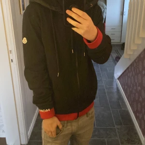 Svart hoodie från Moncler - Säljer en svart hoodie från Moncler med dragkedja och huva. Jackan har långa ärmar och ribbade muddar i rött och blått. Perfekt för en stilren look.