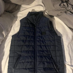 Mörkblå Jack and Jones väst - Snygg mörkblå quiltad väst med dragkedja framtill och två fickor med dragkedjor. Perfekt för lager-på-lager under kyligare dagar. Stilren och enkel design som funkar till det mesta.