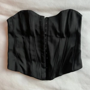 Svart korsett från Zara - Snygg svart korsett från Zara med hakknäppning framtill. Perfekt för att ge en stilren och elegant look. Tillverkad i ett glansigt material som ger en lyxig känsla.