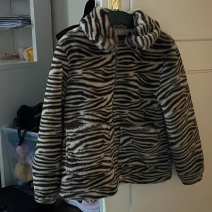 Zebramönstrad pälsjacka med huva - Säljer en cool zebramönstrad pälsjacka med huva. Jackan har långa ärmar och en mysig känsla, perfekt för att sticka ut i mängden. Den är både trendig och bekväm, idealisk för kyligare dagar.