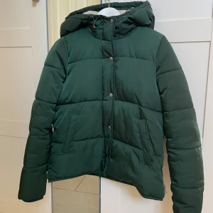 Grön dunjacka med huva - Snygg grön dunjacka med huva och dragkedja. Jackan har en pufferdesign och knappar framtill. Perfekt för kyliga dagar.