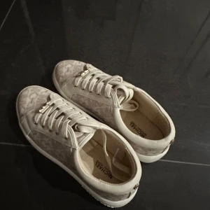 Beige sneakers från Michael Kors - Snygga beige sneakers från Michael Kors med diskret mönster och gulddetaljer. De är onanvändna och i storlek 36, dock för stor i storleken och passar snarare 37-38. 