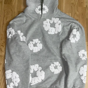 Grå hoodie med blommönster från Denim Tears - Säljer en grå hoodie från Denim Tears med ett unikt vitt blommönster. Tröjan har en klassisk passform med långa ärmar och en bekväm huva. Perfekt för en avslappnad stil.