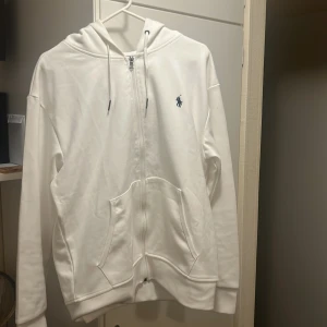 Vit hoodie från Polo Ralph Lauren - Säljer en stilren vit hoodie från Polo Ralph Lauren med dragkedja och det klassiska logotypbroderiet på bröstet. Tröjan har en bekväm passform med långa ärmar och en praktisk huva med dragsko. Perfekt för en avslappnad look.