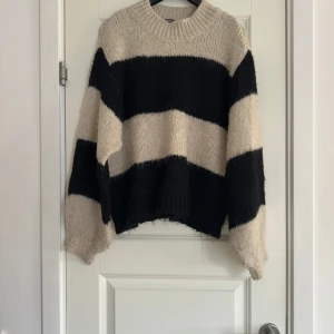 Randig stickad tröja - Säljer en mysig stickad tröja med breda svarta och beige ränder. Tröjan har en avslappnad passform och långa ärmar, perfekt för kyligare dagar. Den är både stilren och bekväm, ett måste i garderoben!