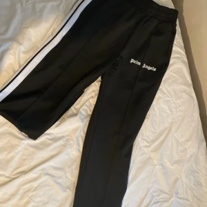 Svarta byxor från Palm Angels - Snygga svarta byxor från Palm Angels med vita sidoremsor. Byxorna har en elastisk midja och är perfekta för en avslappnad stil. Märkesloggan är broderad på sidan. Helt oanvända och lappen sitter kvar, har resten av dressen (dvs tröjan till byxorna) om man skrollar i min profil 