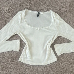 Vit ribbad topp från H&M Divided - Säljer en söt vit ribbad topp från H&M Divided. Toppen har långa ärmar och en fin spetsdetalj runt halsringningen samt en liten rosett i mitten. Knappt använd och i mycket bra skick !!💖💖