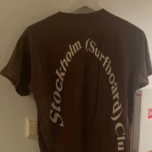 Brun t-shirt med tryck - Säljer en brun t-shirt med texten 'Stockholm (Surfboard) Club' i vitt på baksidan. T-shirten har en klassisk rund hals och korta ärmar. Perfekt för en avslappnad stil.