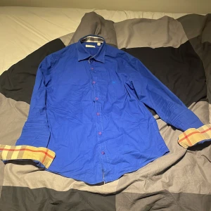 Burberry skjorta - Snygg blå skjorta från Burberry Brit med klassiskt rutigt mönster på insidan av ärmsluten. Skjortan har långa ärmar och knappar framtill. Perfekt för en stilren look.
