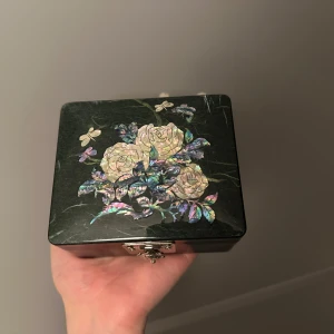 Svart smyckeskrin med blommönster - Elegant svart smyckeskrin med vackert blommönster på locket. Skrinet har en metallås med en sköldpaddsdetalj och små dekorativa fötter. Perfekt för att förvara dina smycken eller andra små skatter.