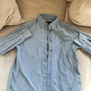 Blå skjorta från Ralph Lauren - Säljer en klassisk blå skjorta från Ralph Lauren. Skjortan har långa ärmar och knappar framtill samt på kragen. Endast använd någon enstaka gång. Junior L ca 14-16 år. 