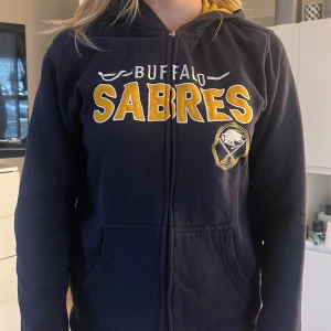 Mörkblå hoodie med Buffalo Sabres-tryck - Snygg mörkblå hoodie med dragkedja och Buffalo Sabres-tryck i gult och vitt. Insidan av huvan är gul, vilket ger en cool kontrast. Perfekt för sportfantaster! Tröjan är från nhl och det står ingen storlek men passar som en S