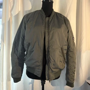 Grön bomberjacka från Hilfiger Denim - Säljer en snygg grön bomberjacka från Hilfiger Denim. Jackan har en klassisk bomberstil med dragkedja framtill och ribbade muddar vid ärmslut och nederkant. Perfekt för en stilren look.