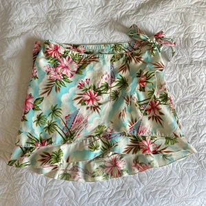 Blommig kortkjol med omlott  - Säljer en superfin blommig kortkjol med tropiskt mönster i rosa, grönt och blått. Kjolen har en söt volangdetalj och knytning i sidan. Perfekt för sommarens varma dagar! Från vintage bikbok