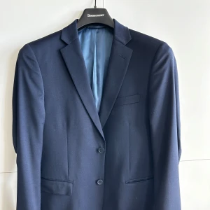 Mörkblå kavaj från Dressmann - Säljer en stilren mörkblå kavaj från Dressmann. Kavajen har en klassisk design med två knappar framtill och en elegant ljusblå insida med röda detaljer.  Storlek 50, slim fit