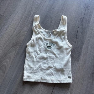 Vit ribbad linnetopp från Pull&Bear - Säljer en vit ribbad linnetopp från Pull&Bear med texten 'San Diego 1993' tryckt på framsidan. Perfekt för en avslappnad stil. Tillverkad i Portugal. Använd 2 ggr