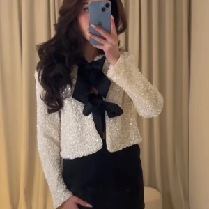 Vit paljettjacka med svart rosett - Snygg vit jacka täckt med paljetter och en stor svart rosett framtill. Jackan har långa ärmar och en croppad passform, perfekt för att sticka ut på festen.