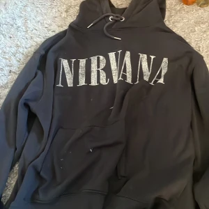 Svart Nirvana hoodie - Säljer en svart hoodie med Nirvana-tryck på framsidan. Den har en klassisk design med huva och dragsko samt långa ärmar. Perfekt för fans av bandet som vill ha en bekväm och stilren tröja.