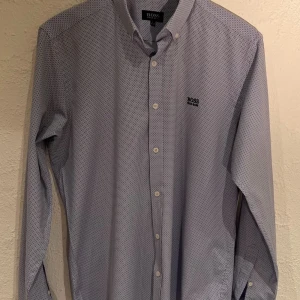 Hugo Boss  ljusblå skjorta strl. L  - Ljusblå skjorta med ett diskret mönster och Hugo Boss logotyp broderad på bröstet. Slim-fit design med en aning stretch för att ge ultimat komfort.