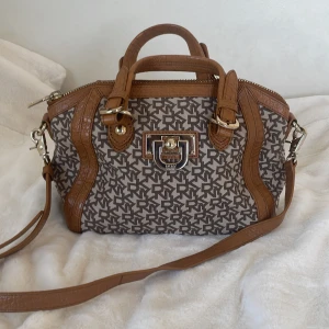 Brun beige handväska från DKNY - Snygg handväska från DKNY i beige med bruna läderdetaljer. Väskan har ett monogrammönster och en praktisk dragkedja. Insidan är fodrad med DKNY-logotyp och har flera fack för enkel organisering. Perfekt för att ge din outfit en stilfull touch!