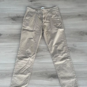 Beige chinos från H&M - Säljer ett par stilrena beige chinos från H&M i slim fit. Byxorna har en klassisk design med knappar och dragkedja framtill. Perfekta för en snygg och avslappnad look.