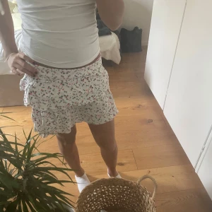 Blommig volangkjol - Söt kortkjol med blommigt mönster i blått och rött på vit bakgrund. Kjolen har volanger som ger en lekfull känsla. Perfekt för varma dagar. storleken är 158