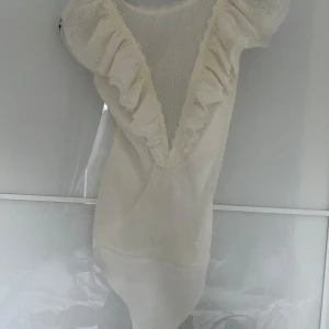 Vit ärmlös body från Bershka - Säljer en söt vit body från Bershka med volangdetaljer och djup v-ringning. Den är ärmlös och har ett diskret prickigt mönster. Perfekt för en stilren look. Passar bra till både kjol och byxor.