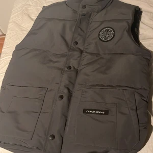 Grå dunväst från Canada Goose - Säljer en stilren grå dunväst från Canada Goose med broderad logga på bröstet. Västen har knappar framtill och praktiska fickor. Perfekt för kyliga dagar, men även varmare dagar. Pris ej hugget i sten. Skriv vid funderingar. Storlek S men sitter även bra på M