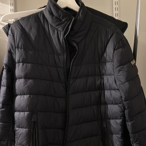 Svart pufferjacka från Napapijri - Säljer en stilren svart pufferjacka från Napapijri. Jackan har en quiltad design med dragkedja framtill och två praktiska fickor. Perfekt för kyligare dagar med sin varma och bekväma passform.