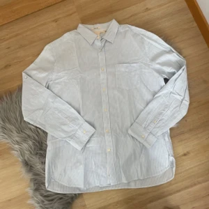 Ljusblå och vit randig skjorta från H&M - Säljer en stilren ljusblå och vit randig skjorta från H&M. Skjortan har långa ärmar och knappar framtill. Perfekt för en avslappnad men ändå snygg look. Tillverkad i mjuk bomull för extra komfort.
