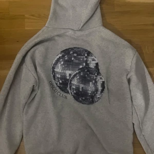 Grå hoodie med discokulor - Säljer en grå hoodie med tryck av två discokulor och texten 'Disco Club' på ryggen. Perfekt för en avslappnad stil med en touch av festkänsla. Hoodien har långa ärmar och en klassisk passform.