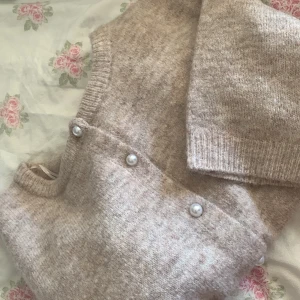 Beige kofta med pärldetaljer från 157 - Säljer en mysig beige kofta från 157 med fina pärldetaljer längs knäppningen. Koftan har korta ärmar och en mjuk, stickad textur. Använd 1 gång