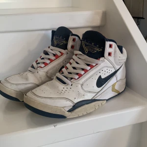 Nike Air sneakers i vitt och blått - Klassiska Nike Air sneakers i vitt med blå och röda detaljer. Skorna har en hög design med snörning och perforerade detaljer för ventilation. Den ikoniska Nike-loggan syns på sidan och hälen.