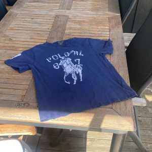 Blå t-shirt från Ralph Lauren - Säljer en blå t-shirt från Ralph Lauren med ett stort tryck av en polospelare och texten 'Polo 67' på framsidan. T-shirten har korta ärmar med siffran '3' tryckt på ena ärmen. Perfekt för en avslappnad stil!