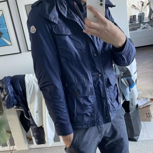 Blå jacka från Moncler - Säljer en snygg blå jacka från Moncler med flera fickor och knappar. Jackan har en huva och Moncler-loggan på ärmen. Perfekt för en stilren look. Nypris ligger runt 13000kr 