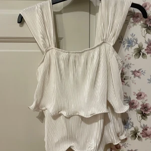 Vit topp - Säljer en elegant vit plisserad topp med breda axelband och volangdetaljer. Perfekt för en stilren look. Fint skick 