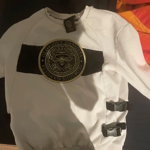 Vit sweatshirt från Glorious Gangsta - Snygg vit sweatshirt från Glorious Gangsta med svart panel och guldtryck på bröstet. Tröjan har ribbade muddar och unika spänndetaljer på sidan. Perfekt för en stilren och modern look.