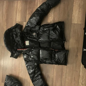 Svart dunjacka från Moncler - Säljer en snygg svart dunjacka från Moncler. Jackan har en glansig finish och en avtagbar huva. Perfekt för kyliga dagar med sin pufferdesign som håller dig varm. Den har en dragkedja framtill och en klassisk Moncler-logga på bröstet.