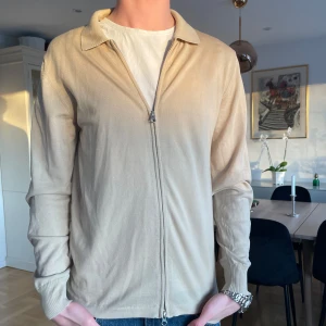 Beige tröja med dragkedja - Säljer en stilren beige J.Lindeberg tröja med dragkedja framtill. Perfekt för både vardag och mer uppklädda tillfällen. Tröjan har långa ärmar och en klassisk krage.