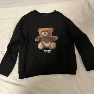 Svart tröja med nalletryck från Moschino - Säljer en svart tröja från Moschino med ett stort nalletryck på framsidan. Tröjan har långa ärmar och är tillverkad i bomull för en bekväm passform. Perfekt för dig som vill ha en stilren men lekfull look.