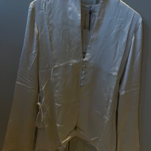Krämfärgad blus från Avora - Helt ny blus från Avora! Blusen har långa ärmar och en stilren design med knappar framtill och ett knytband i midjan. Perfekt för en chic och sofistikerad look. Den är i färgen ”cream”