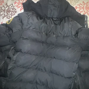 Svart pufferjacka från Zavetti Canada - Säljer en svart pufferjacka från Zavetti Canada med en broderad logga på ärmen. Jackan har en dragkedja framtill och är perfekt för kalla dagar. Den har en hög krage och är långärmad för extra värme.