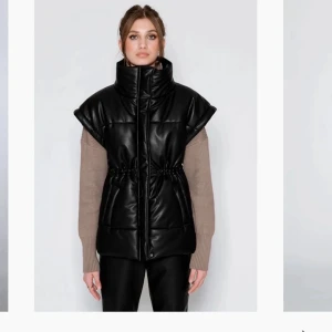 Väst i skinnimitation med hög krage - Snygg svart väst med hög krage och korta ärmar. Västen har en dragkedja framtill och elastisk midja för en bekväm passform. Perfekt för lager-på-lager under kyliga dagar. I skinnimitation från märket LXA. Väl använd men i gott skick.