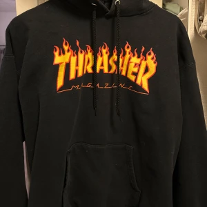 Svart hoodie från Thrasher - Säljer en svart hoodie från Thrasher med det ikoniska flammande logotyptrycket i rött och gult på framsidan. Perfekt för en avslappnad stil med en justerbar huva. Passar dig som är 170-187cm lång! 