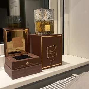 Khamrah Eau de Parfum från Lattafa - Elegant parfymflaska med ett lyxigt mönstrat lock och gyllene vätska. Förpackningen är brun med guldtext och innehåller 100 ml av doften Khamrah. Perfekt för den som söker en sofistikerad och unik doftupplevelse. 96/100 ml kvar 