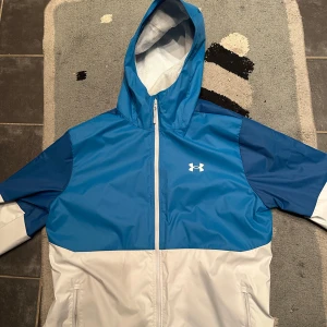 Blå och vit vindjacka från Under Armour - Säljer en snygg blå och vit vindjacka från Under Armour. Jackan har en dragkedja framtill och en praktisk huva. Perfekt för blåsiga dagar och sportiga aktiviteter. Den har långa ärmar och är tillverkad i ett lätt syntetmaterial. Jackan har även aldrig använts.